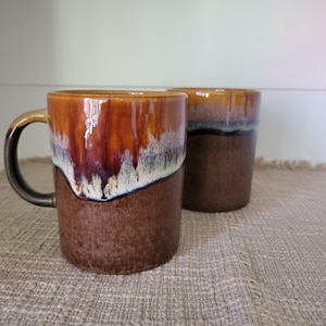 May include: Two ceramic mugs featuring a two-tone glaze. The upper portion has a glossy, dripping finish in shades of amber, cream, and dark brown, while the lower section is a solid, matte-textured dark brown. Each mug has a sturdy handle.