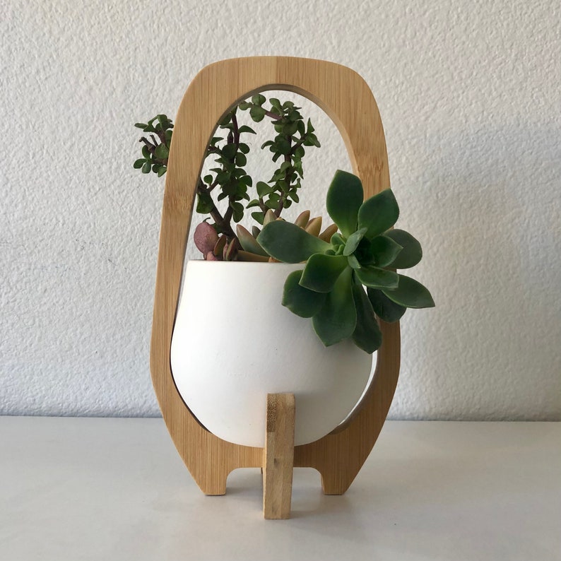 May include: A white ceramic planter bowl held within a light-toned bamboo wood frame. The frame features an arched top and a central vertical support base. Inside the planter are several green succulent plants with varied leaf shapes and textures.