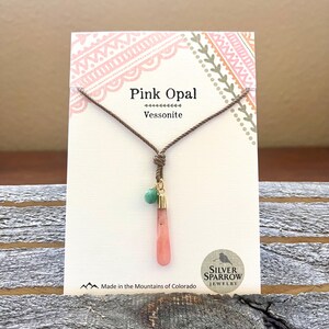 May include: A pink opal gemstone pendant necklace with a green gemstone accent bead. The necklace is made with brown cord and gold accents. The card reads "Pink Opal Vessonite" and "Silver Sparrow Jewellery".