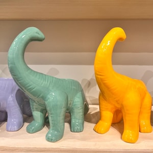 Puede incluir: Una exhibición de macetas de dinosaurios de cerámica en varios colores. La imagen presenta una maceta de dinosaurio verde claro, amarilla y morada. Las macetas tienen cuellos largos y están sobre una estantería de madera. Los dinosaurios están esmaltados y tienen un acabado brillante.