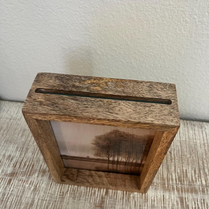 May include: A rectangular wooden frame with a natural grain finish featuring a horizontal slot at the top. The frame contains a printed image of a landscape with dark trees against a soft, light-colored background, set within the wooden structure.