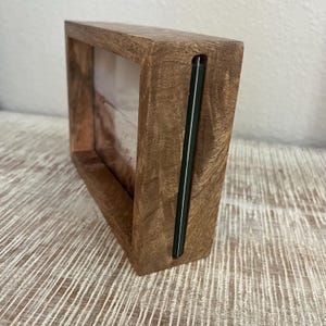 May include: A rectangular wooden picture frame with a thick, natural grain finish. The frame features a side-loading slot design with a thin, dark green edge visible along the vertical opening where a photograph or print can be inserted into the center.