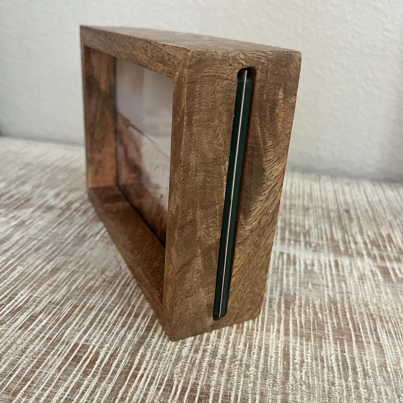 May include: A rectangular wooden picture frame with a thick, natural grain finish. The frame features a side-loading slot design with a thin, dark green edge visible along the vertical opening where a photograph or print can be inserted into the center.