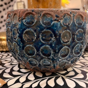 May include: A blue and brown ceramic planter with a textured, circular pattern. The planter is round and has a wide opening.