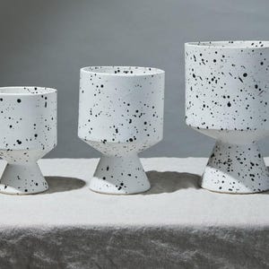 Könnte beinhalten: Drei weiße Keramik-Pflanzgefäße mit schwarzen Sprenkeln. Jedes Pflanzgefäß hat eine zylindrische Oberseite und einen geometrischen Sockel. Die Pflanzgefäße sind unterschiedlich groß und bilden eine gestaffelte Anordnung. Ideal für Pflanzen im Innen- oder Außenbereich.
