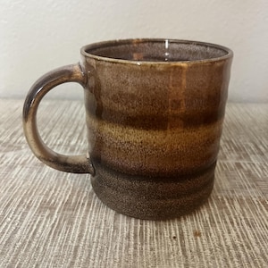 May include: A ceramic mug featuring a multi-toned glaze with horizontal bands of dark brown, golden tan, and a lighter beige at the rim. The mug has a smooth, rounded handle and a cylindrical body with a slightly textured, speckled finish.