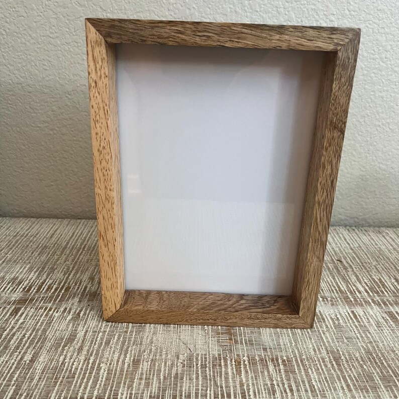 May include: A rectangular picture frame constructed from light brown wood with a visible natural grain pattern. The frame features a deep profile and surrounds a blank, white rectangular insert, providing a clean and neutral display area.
