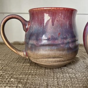 May include: A ceramic mug featuring a rounded body and a curved handle. The glaze displays a gradient of colors, transitioning from a deep red at the rim to a muted purple in the center, and ending in a light tan at the base.