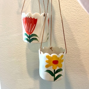Puede incluir: Dos macetas colgantes de cerámica con bordes festoneados. Una maceta presenta un diseño de tulipán rojo con hojas verdes, y la otra tiene una flor amarilla con un centro rojo y hojas verdes. Las macetas cuelgan de correas de cuero marrón.