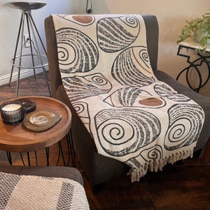May include: A cream-colored throw blanket with a black and brown seashell pattern. The blanket is draped over a chair with a brown fabric upholstery.