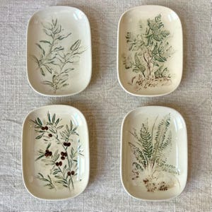 May include: Four off-white rectangular ceramic dishes with botanical designs. Each dish features a different green and brown plant illustration, including ferns and branches with berries. The dishes are arranged in a square formation.