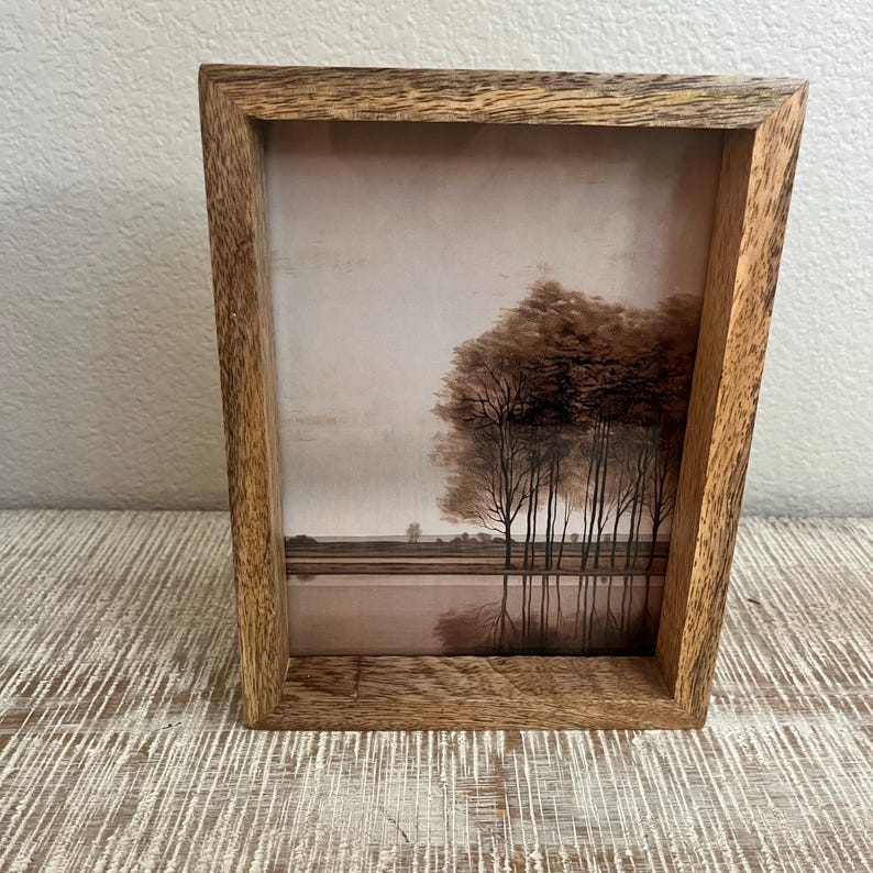 May include: A rectangular wooden picture frame with a natural grain finish containing a sepia-toned print of a landscape. The image depicts a line of trees along a horizon with a reflection visible in the water below, set against a pale sky.