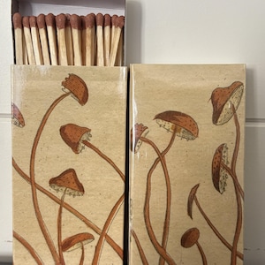 May include: A matchbox set with a beige background and a repeating illustration of brown mushrooms. The matchbox is open, revealing a row of wooden matches with brown tips. The design is repeated on the matchbox cover.