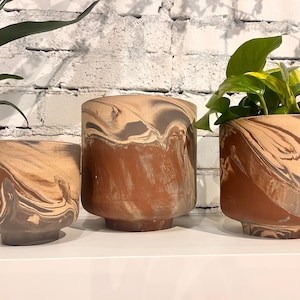May include: Three decorative planters with a marbled design in shades of brown, tan, and grey. The planters are cylindrical with a slightly flared base. One planter holds a green plant. The planters are arranged on a white surface.