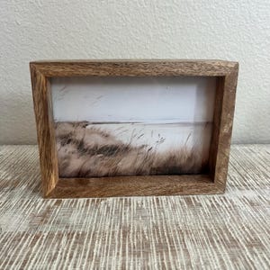 May include: A rectangular wooden picture frame with a natural brown grain finish. The frame contains a print depicting a coastal landscape with tall, thin grasses in the foreground and a calm, light-colored horizon line in the background.