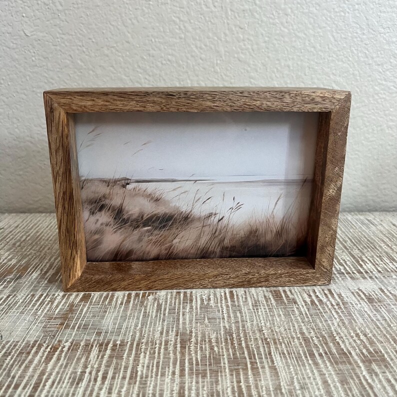May include: A rectangular wooden picture frame with a natural brown grain finish. The frame contains a print depicting a coastal landscape with tall, thin grasses in the foreground and a calm, light-colored horizon line in the background.