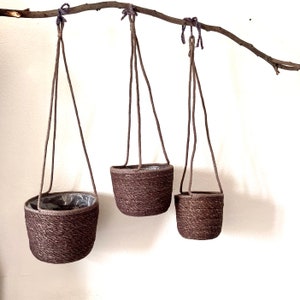 Puede incluir: Tres macetas colgantes marrones con cuerdas. Las macetas están hechas de un material tejido y cuelgan de una rama.