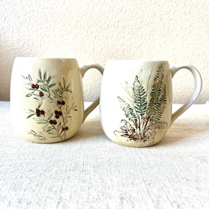 May include: Two ceramic mugs with cream-colored glaze. One mug features a botanical design of green leaves and red berries. The other mug has a fern design in shades of green and brown. Both mugs have curved handles.