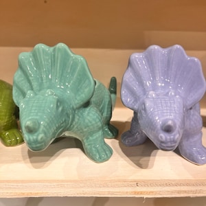 May include: Ceramic triceratops figurines in shades of green and lavender. The dinosaur sculptures have detailed textured skin and are posed in a crouching position. The figurines are displayed on a light-coloured wooden shelf.