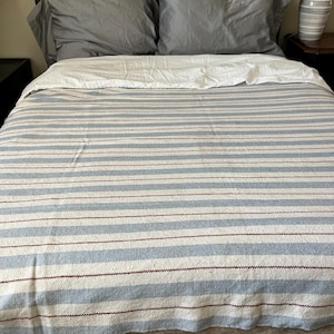 May include: A woven bedspread featuring a pattern of horizontal stripes in light blue, off-white, and thin dark brown lines. The textile covers a bed with a white flat sheet folded over the top edge and two grey pillows positioned at the head.