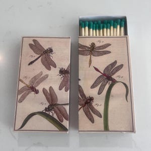 May include: Two decorative matchboxes with dragonfly illustrations. One box is open, revealing green-tipped matches. The boxes feature a vintage design with dragonfly drawings in shades of brown and beige, with green accents.