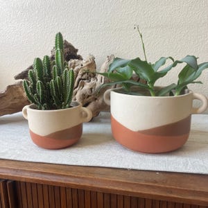 May include: Two ceramic planters with a cream, brown, and terracotta color scheme. One planter holds a cactus, the other a leafy green plant. Both have small handles and sit on a neutral-colored surface.