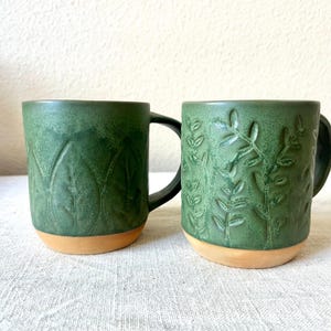 May include: Two green ceramic mugs with leaf and plant designs. The mugs have a dark green exterior with a lighter tan base. One mug features a large leaf pattern, while the other has a detailed plant design. Both have black handles.