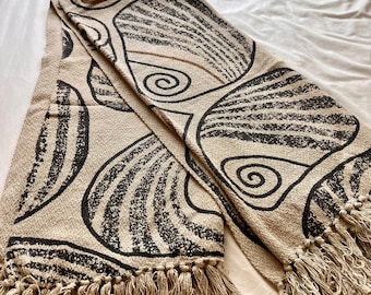 May include: A beige throw blanket with a black and brown abstract pattern. The blanket has a fringe border.