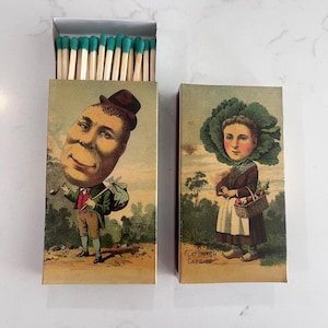 Puede incluir: Dos cajas de cerillas vintage con ilustraciones. Una presenta a un hombre con una cara grande, el texto "A Potato Faced Man". La otra representa a una mujer con cabeza de col, con el texto "Flat Dutch Cabbage". Las cerillas tienen puntas turquesas.