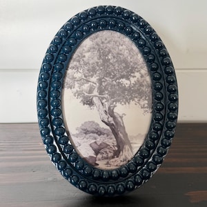 May include: Oval navy blue picture frame with a beaded border. The frame holds a black and white print of a tree and landscape. The frame is displayed on a dark wooden surface.
