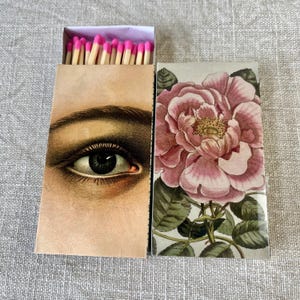 Puede incluir: Dos cajas de cerillas decorativas. Una presenta una ilustración detallada de un ojo, la otra una rosa rosa con hojas verdes. La caja de cerillas abierta a la izquierda revela cerillas con punta rosa.