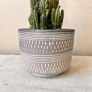 May include: A gray ceramic planter with a white geometric pattern. The planter has a wide, round base and a slightly flared rim. A cactus plant is growing in the planter.