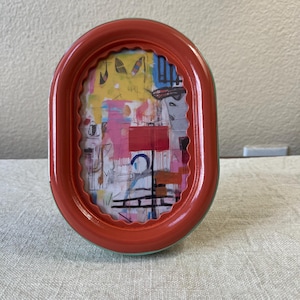 Può includere: Cornice ovale color corallo con bordo interno verde chiaro, che mostra arte astratta colorata. L'opera d'arte presenta tonalità rosa, gialle e blu. La cornice è su una superficie neutra. La forma della cornice è ovale.