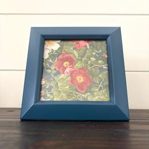 Peut inclure: Un cadre photo carré avec une finition bleu marine mate et lisse. Le cadre contient une impression botanique représentant des fleurs rouges et blanches éclatantes aux centres jaunes, entourées de feuilles vertes détaillées sur fond bleu.