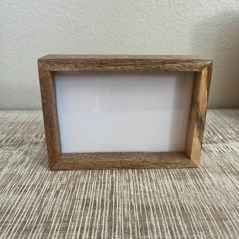 May include: A rectangular picture frame constructed from natural brown wood with a visible grain pattern. The frame features a thick, sturdy border surrounding a blank, white rectangular center space, designed for displaying photographs or artwork.