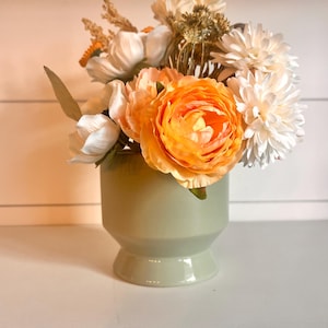 May include: A floral arrangement in a light green ceramic vase. The bouquet features large orange and white flowers, with smaller white and beige blooms. The vase has a pedestal base and a rounded body, creating a modern aesthetic.