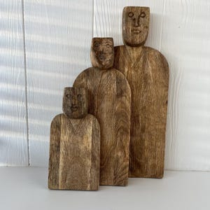 May include: Three wooden sculptures of stylized figures with carved faces and simple clothing. The figures vary in size, with the largest standing approximately 12 inches tall. The wood grain is visible, giving each piece a natural, rustic appearance.