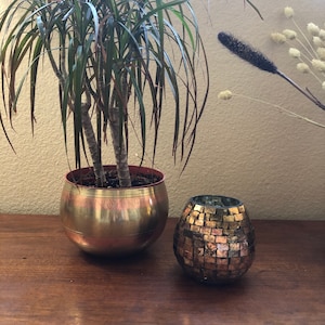 Peut inclure: Un pot en métal doré avec une plante verte à l'intérieur. Le pot a un motif décoratif. Un petit bougeoir en mosaïque dorée est posé sur une surface en bois.