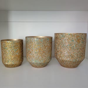 Puede incluir: Un conjunto de tres macetas cilíndricas con un exterior texturizado que combina láminas de pan de oro con pequeños fragmentos de piedra turquesa y blanca. Cada maceta tiene un borde interior dorado y una base cónica.