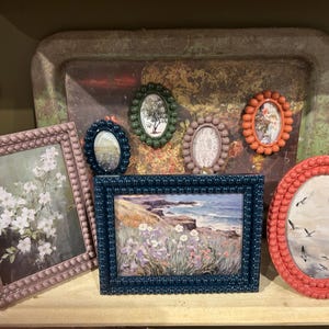 Könnte beinhalten: Eine Sammlung gerahmter Kunstwerke in verschiedenen Formen und Größen. Die Rahmen sind in Rosa-, Blau- und Orangetönen gehalten und haben ein strukturiertes, perlenbesetztes Design. Die Kunstwerke zeigen Blumenarrangements, Landschaften und Vögel.