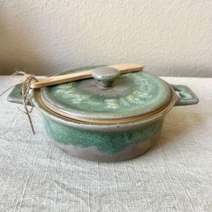 May include: A small, round, green ceramic casserole dish with a lid and a wooden spoon. The dish has a handle on each side and a knob on the lid. The lid has embossed text. A twine bow is tied to the handle.