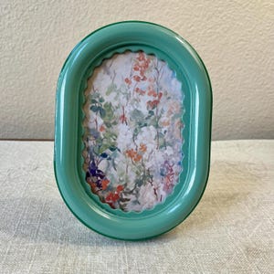 May include: Oval-shaped, mint green picture frame with a scalloped edge. The frame holds a floral print with red, white, and green flowers. The frame is made of a glossy material and sits on a neutral-colored surface.
