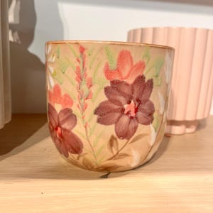 Puede incluir: Una maceta de cerámica beige con un diseño floral que presenta flores burdeos, coral y blancas. La maceta tiene una forma redondeada y está decorada con hojas y tallos verdes. En el fondo se ve una maceta rosa.