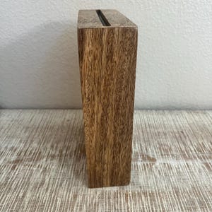 May include: A rectangular wooden block stand featuring a natural brown wood grain pattern and a narrow, centered vertical slot running along the top edge, designed to hold thin items upright in a stable, upright position.