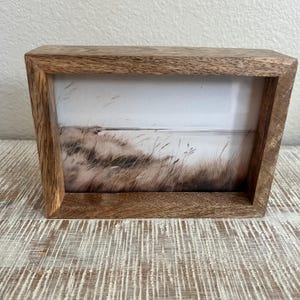 May include: A rectangular wooden picture frame with a natural brown grain finish. The frame contains a print depicting a landscape scene with tall, thin grasses blowing in the wind against a light, muted sky and a distant horizon line.