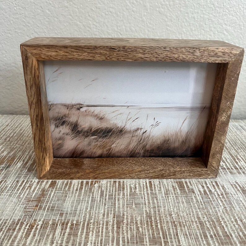 May include: A rectangular wooden picture frame with a natural brown grain finish. The frame contains a print depicting a landscape scene with tall, thin grasses blowing in the wind against a light, muted sky and a distant horizon line.