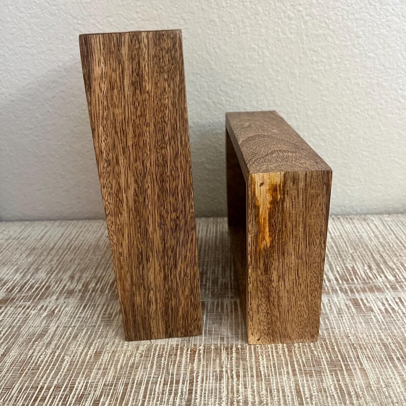 May include: Two rectangular wooden blocks with a visible brown wood grain pattern. The taller block stands vertically on its side, while the shorter block is positioned next to it, displaying a natural wood texture and a light-colored knot.