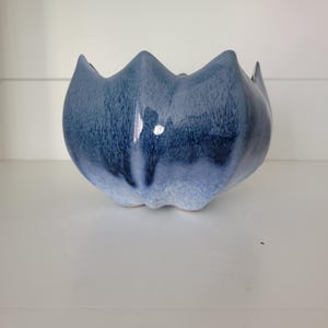 Peut inclure: Un bol en céramique avec un bord festonné en forme de pétale et un vernis brillant qui passe d'un bleu marine profond en haut à un bleu ciel et blanc moucheté plus clair près de la base, présentant une forme courbe, lisse et organique.