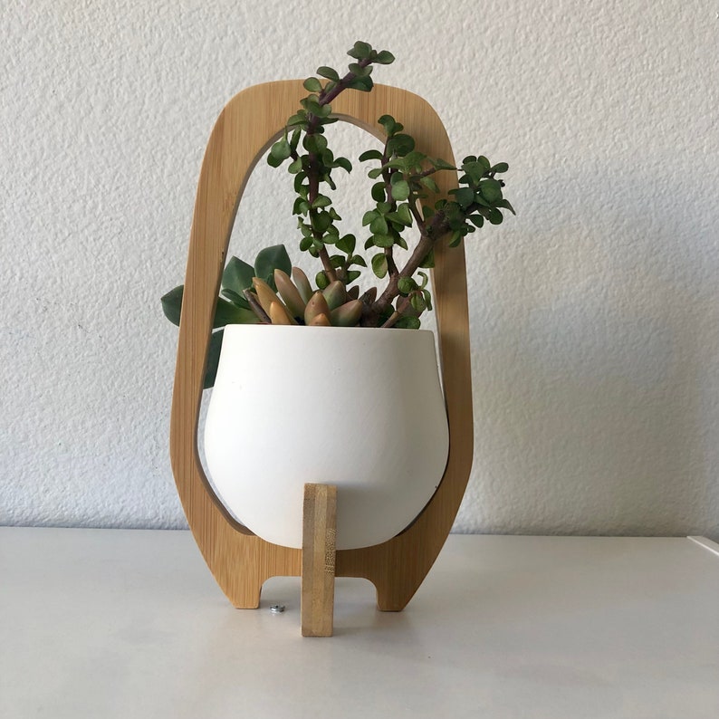 May include: A white ceramic planter bowl held within a light-toned wooden frame. The planter contains a variety of green and reddish-tinted succulent plants with thick, fleshy leaves. The wooden frame features an arched top and a central support.