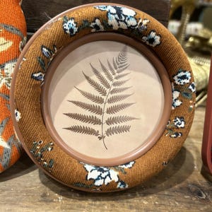May include: Round picture frame with a brown floral fabric border. The frame holds a fern print on a beige background. The frame is sitting on a wooden surface.